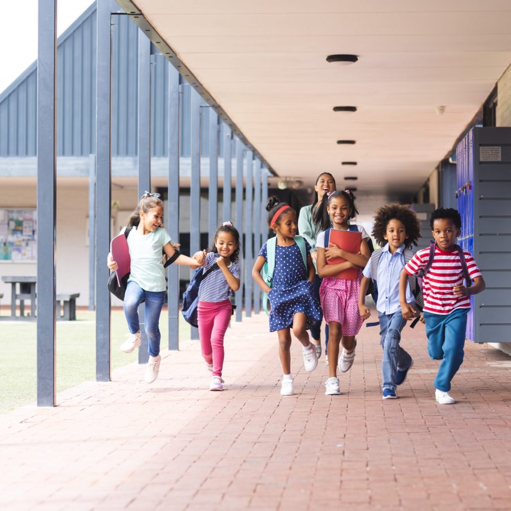 In school, a diverse group of students are running down a hallway outdoors. They are wearing casual clothes and have joyful expressions, unaltered.